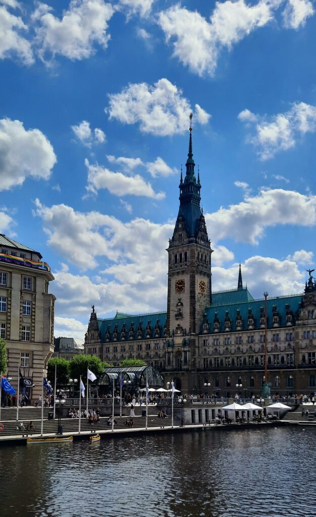 Hamburg Rathaus with its central clock tower, seen across the water beneath a blue sky with scattered clouds.