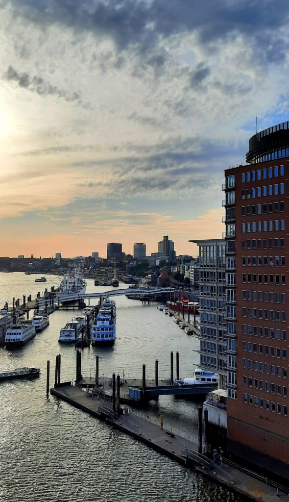 View from the Elbphilharmonie Plaza over Hamburg harbour, with boats, waterfront buildings and the Elbe beneath an evening sky.