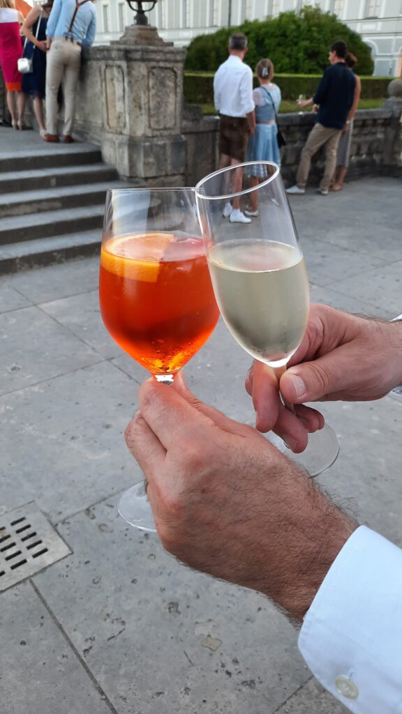 Two glasses raised for a toast outdoors at Nymphenburg Palace in Munich, with other concert visitors in the background.