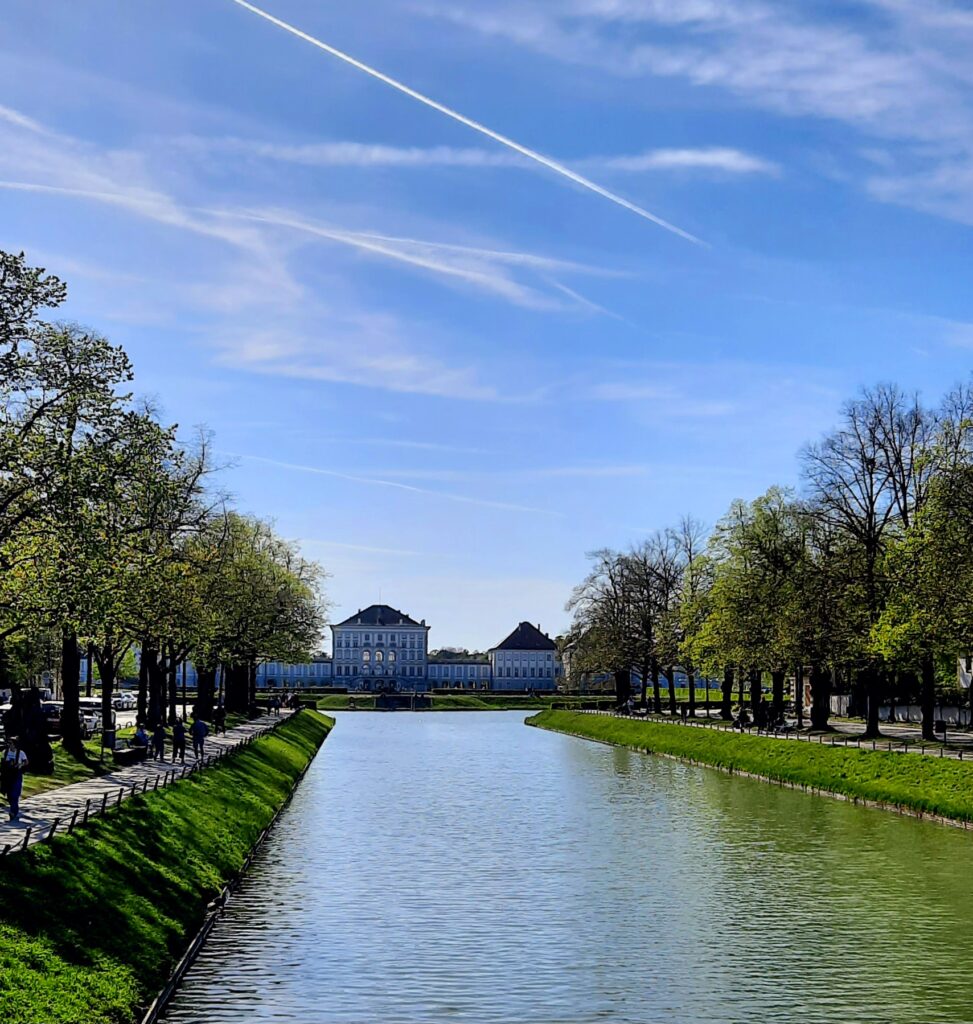 View of Nymphenburg Palace in Munich across the long canal, framed by trees beneath a blue spring sky.