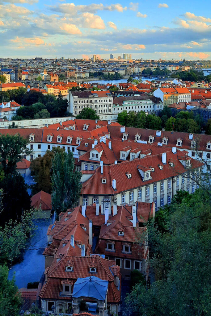 Panoramic view of Prague's red rooftops, historic buildings and the Vltava River beneath a partly cloudy sky.