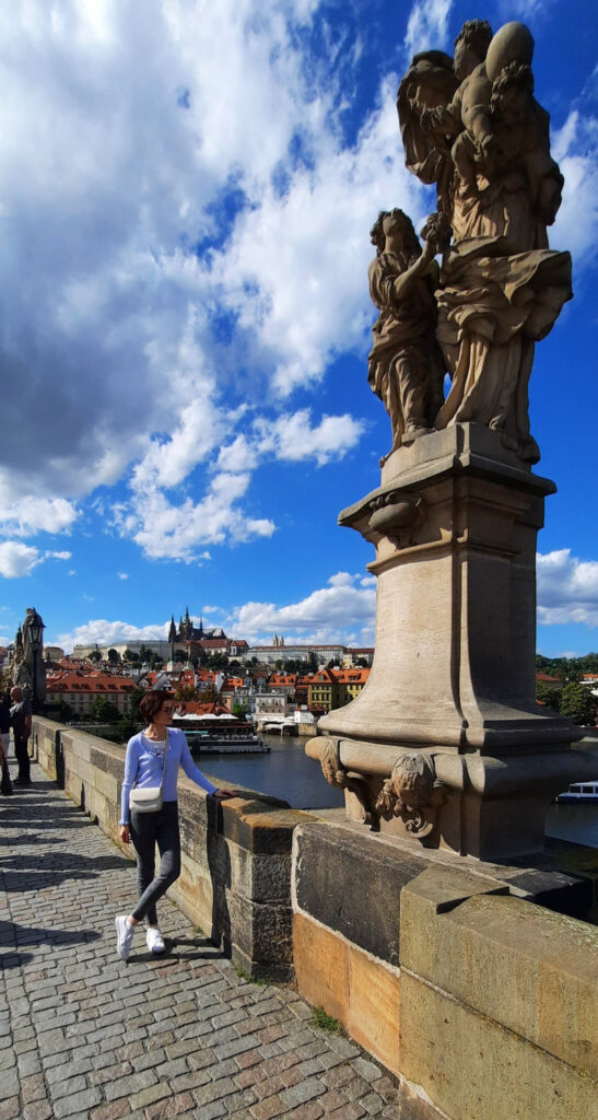 Woman standing beside a stone statue on Charles Bridge in Prague, with the Vltava River and Prague Castle in the background.