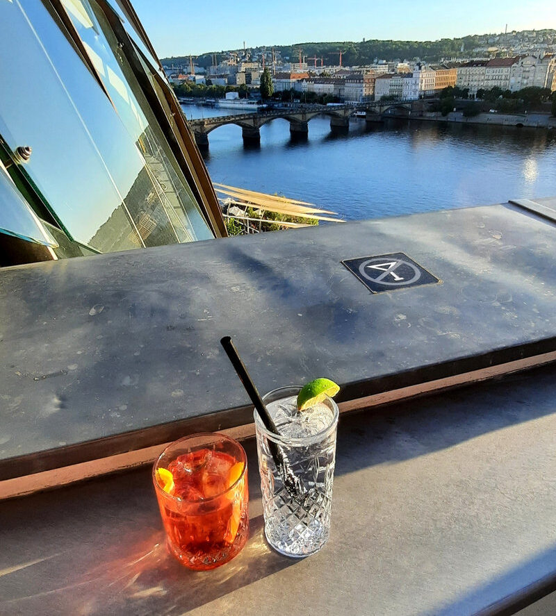 Two drinks at the Dancing House rooftop bar overlooking the Vltava River and Prague's historic buildings.