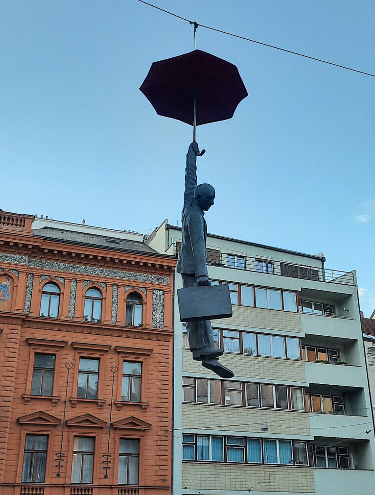 Side view of a suspended figure holding an umbrella and briefcase above a Prague street.