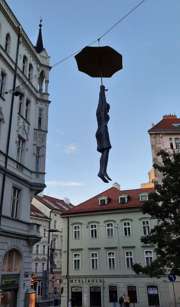 Suspended figure holding an umbrella above a street in Prague, surrounded by historic buildings.