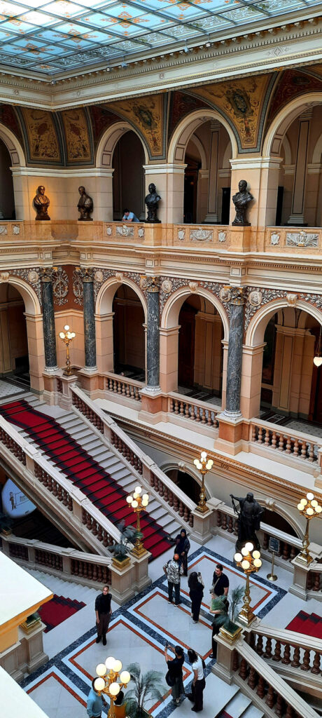 Grand staircase inside Prague's National Museum, with red-carpeted steps, ornate arches, marble columns and visitors exploring the hall.