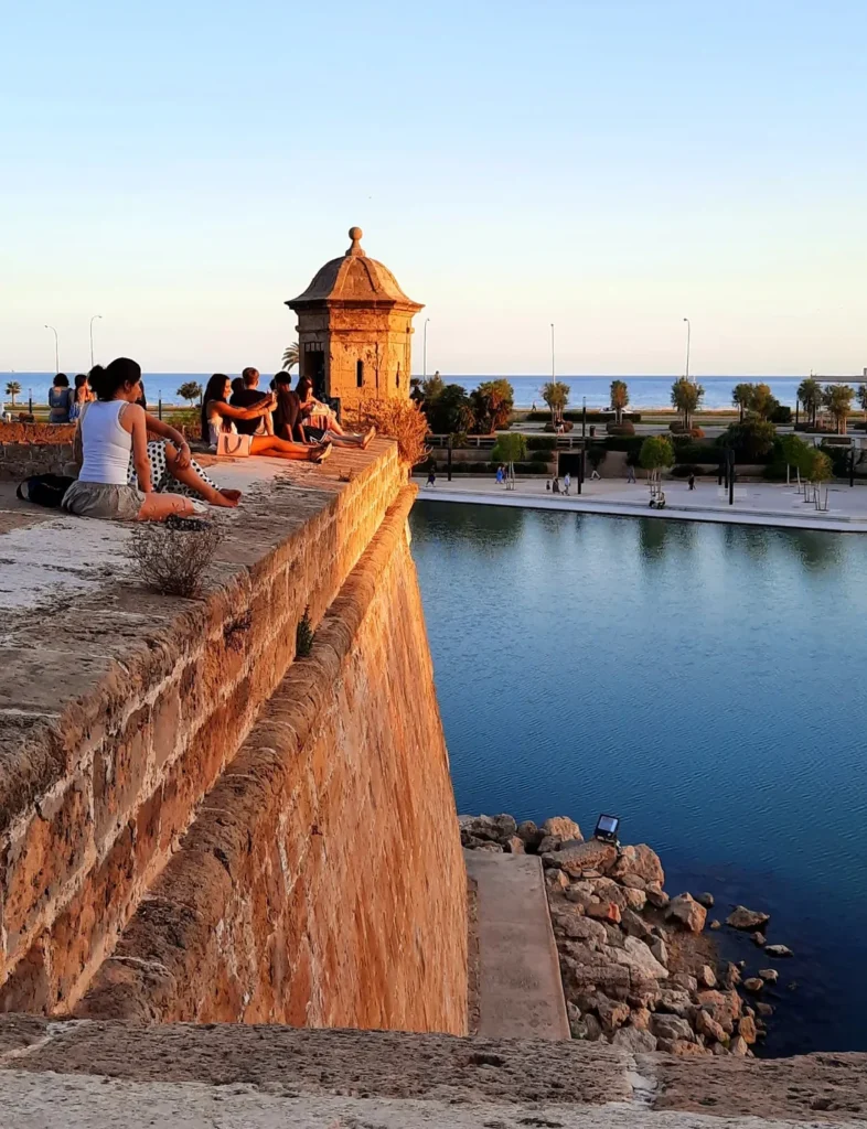 People sitting on historic stone walls overlooking the sea in Palma, Mallorca, at sunset